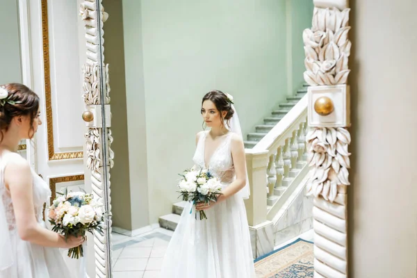 Aşık genç bir çift. Gelinin koyu renk saçları ve uzun bir peçesi var. Gelin buketi. Aynadaki yansıma. Beyaz mermer merdiven.