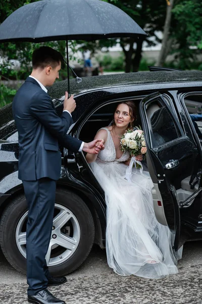 恋爱中的年轻夫妇 新娘有一头乌黑的头发和长长的面纱 新娘花束新郎帮助新娘下了车 手里拿着黑色雨伞 — 图库照片