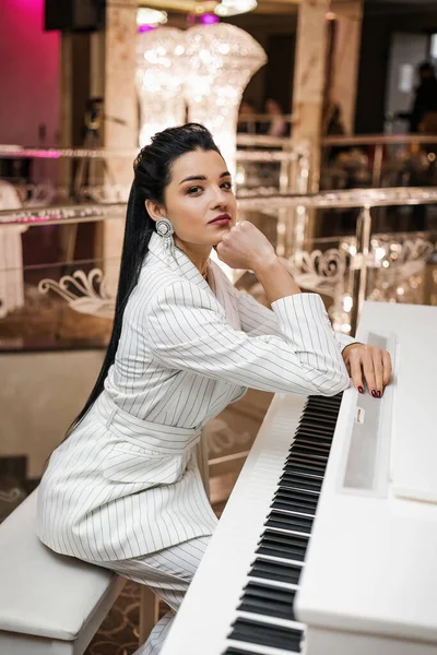 迷人的黑发姑娘穿着白色西服坐在一架白色钢琴前 华丽的气氛奢华的细节 — 图库照片