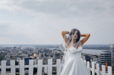 Uzun bir evin çatısında duvağı olan şık, nazik bir gelin. Gökdelenleri ve iş merkezleri olan güzel bir şehir manzarası açılıyor..