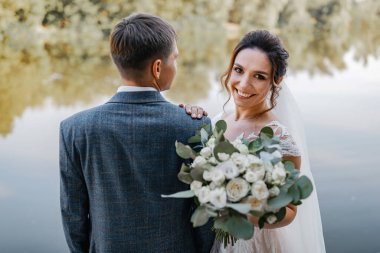The wedding couple smiles and pauses for the camera. A river in the background. The bride has a bridal bouquet