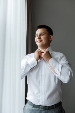 Beyaz gömlekli çekici bir damat pencerenin yanında poz veriyor. Doğal ışık