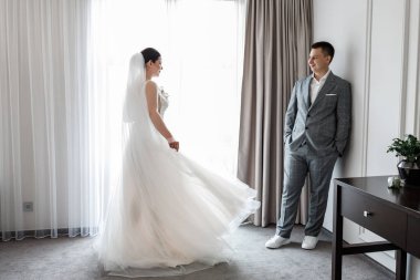 Gri takım elbiseli damat ve pencerenin yanında uzun peçeli gelin..