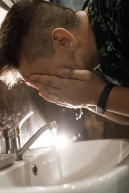 Bir adam yüzünü banyoda yıkar, ışık arkadan yanar..