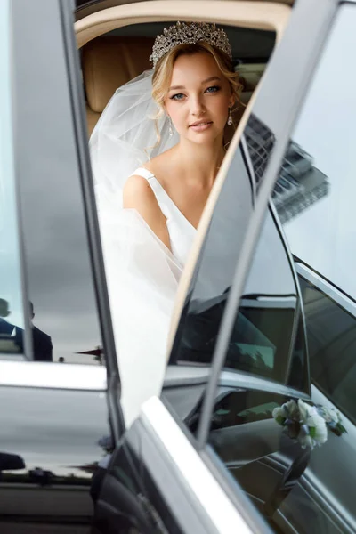Enchanting blonde bride in a wedding dress with a long beautiful veil. In the back seat of a black car.