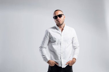 Beyaz gömlekli, siyah gözlüklü şık bir adam beyaz bir arka plana karşı poz veriyor..