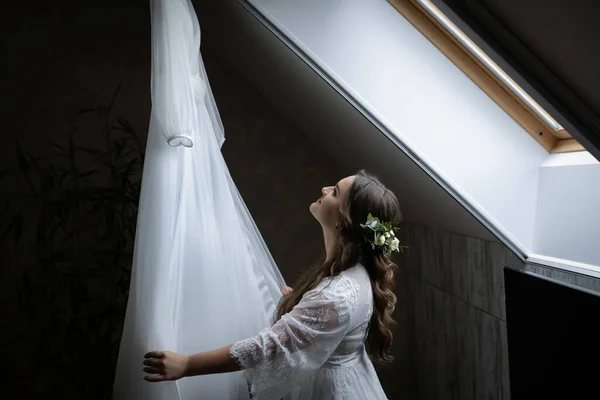 가운을 귀여운 신부님 창가에 옷걸이에는 드레스를 입는다 꽃으로 머리를 아름다운 — 스톡 사진