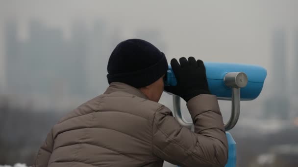 Par une journée d'hiver, un gars regarde à travers les jumelles sur la plate-forme d'observation vers le paysage urbain et le centre d'affaires de Moscou. — Video