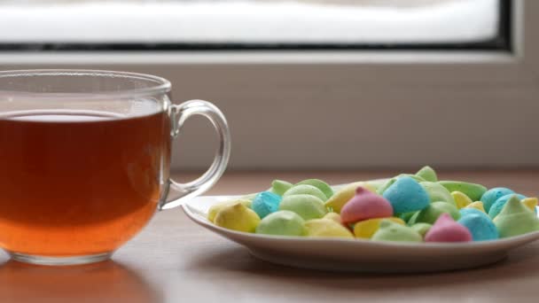 A large mug of tea and colorful sweets in a plate on the windowsill by the window in winter. The camera is in motion. Selective focus — 图库视频影像