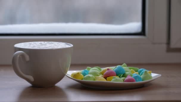 A large mug of coffee with milk or mochacino and colorful sweets in a plate on the windowsill in winter. Selective focus — Wideo stockowe