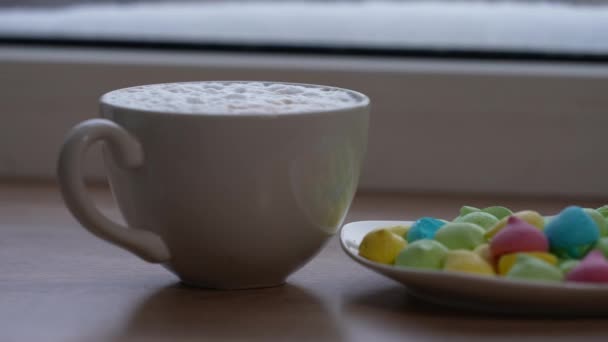 A large mug of coffee with milk or mochacino and colorful sweets in a plate on the windowsill in winter. Selective focus — Vídeo de Stock