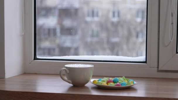 A large mug of coffee with milk or mochacino and colorful sweets in a plate on the windowsill in winter. Selective focus — Stok video