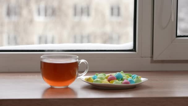 Una taza grande de té y dulces de colores en un plato en el alféizar de la ventana en invierno. La cámara está en movimiento. Enfoque selectivo — Vídeo de stock