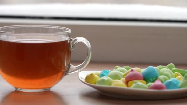 Una taza grande de té y dulces de colores en un plato en el alféizar de la ventana en invierno. La cámara está en movimiento. Enfoque selectivo — Vídeo de stock