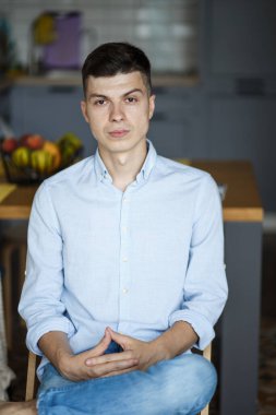 Mutfakta sandalyede oturan ve gülümseyen genç bir adamın portresi. Kot pantolon ve mavi gömlek giymişti. Ellerini sandalyede tutuyor..