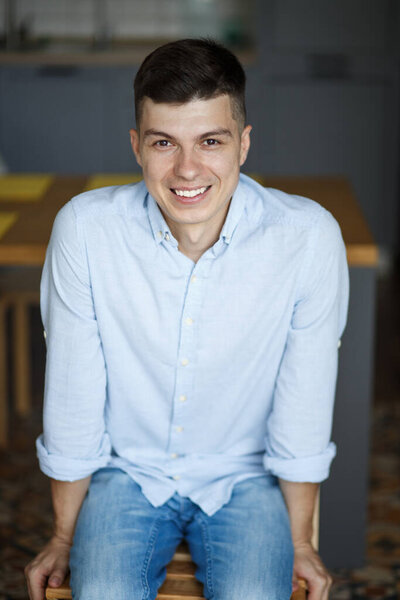 portrait of a young stylish guy who sits on a chair in the kitchen with his arms crossed. dressed in blue jeans and a blue shirt. 