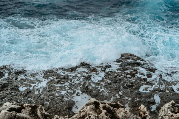 Fırtınadan sonra güçlü dalgalar sıçrıyor. Vahşi kayalık deniz kıyısı, rüzgarlı hava. Arka planda beyaz dalga olan soyut mavi deniz suyu. Okyanus dalgaları çarpıyor. Suyun gücü.