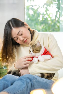 Asyalı kadın gülüyor ve oyun sırasında mutlu ve kedisine kırmızı Noel Baba battaniyesi giyiyor.