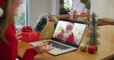 Noel Baba şapkası takan beyaz bir kadın Noel 'de evde dizüstü bilgisayarında bir video görüşmesi yapıyor. Noel zamanı covid 19 salgınında sosyal mesafeler
