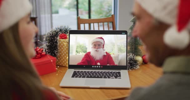 クリスマスの間にラップトップでビデオコールを持っているサンタの帽子をかぶっている白人カップルの後部ビュー クリスマスの時期に流行した19種のパンデミックの間の社会的距離は — ストック動画