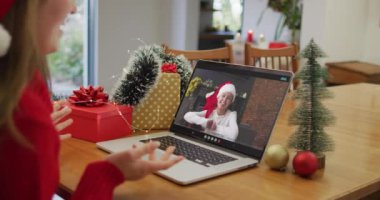 Noel Baba şapkası takan beyaz bir kadın Noel 'de evde dizüstü bilgisayarında bir video görüşmesi yapıyor. Noel zamanı covid 19 salgınında sosyal mesafeler