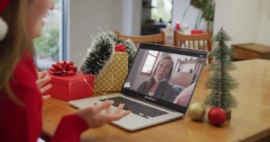Yılbaşı zamanı yüz maskesi takan bir bayanla birlikte laptoptaki mutlu beyaz kadın görüntüsü. Coronavirus covid 19 salgını sırasında Noel, şenlik ve iletişim teknolojisi.