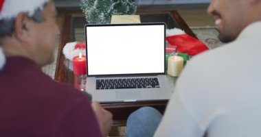 Gülümseyen çift ırklı baba ve yetişkin oğul dizüstü bilgisayar ile noel görüntülerini çekiyorlar. Noel, şenlik ve iletişim teknolojisi.