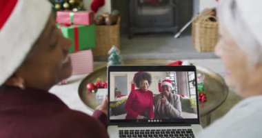 Kıdemli çeşitli bayan arkadaşlar, ekranda gülümseyen bir aileyle yılbaşı videosu için dizüstü bilgisayar kullanıyorlar. Noel, şenlik ve iletişim teknolojisi.