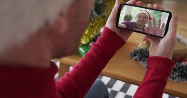 Kafkasyalı bir adam Noel 'de ekranda gülümseyen bir aileyle akıllı telefon kullanıyor. Noel, şenlik ve iletişim teknolojisi.