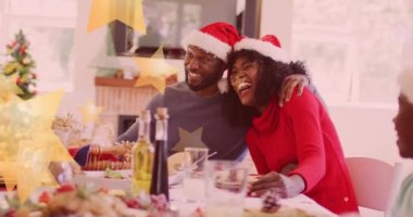 Mutlu Afro-Amerikan ailesinin üzerine düşen yıldızların animasyonu. Noel Baba şapkası takıp yemek yiyorlar. Noel, kış, gelenek ve kutlama konsepti dijital olarak oluşturulmuş video.