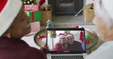 Kıdemli çeşitli bayan arkadaşlar, ekranda gülümseyen bir aileyle yılbaşı videosu için dizüstü bilgisayar kullanıyorlar. Noel, şenlik ve iletişim teknolojisi.