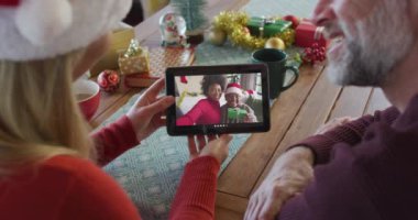 Noel Baba şapkalı, gülümseyen beyaz bir çift ekranda aileleriyle birlikte noel videosu için tablet kullanıyorlar. Noel, şenlik ve iletişim teknolojisi.