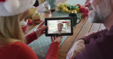 Noel Baba şapkalı, gülümseyen beyaz bir çift ekrandaki adamla noel videosu için tablet kullanıyor. Noel, şenlik ve iletişim teknolojisi.