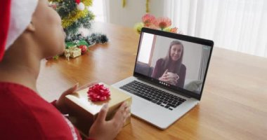 Noel Baba şapkalı Afro-Amerikalı bir kadın, ekrandaki kadınla yılbaşı görüntüsü için laptop kullanıyor. Noel, şenlik ve iletişim teknolojisi.