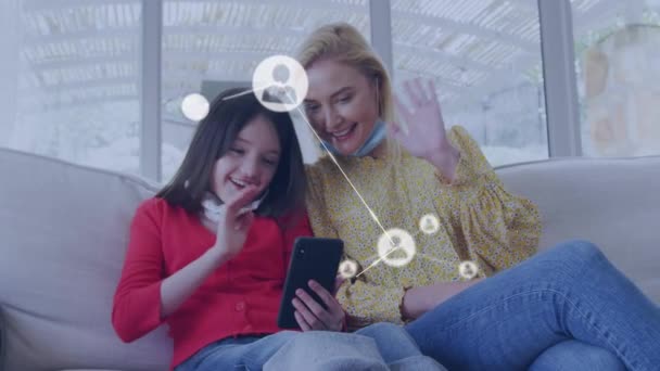 スマートフォンを使用して顔のマスクを持つ白人の母親と娘の上にメディアアイコンのアニメーション 世界的なビジネス デジタルインターフェースをデジタルで生成する — ストック動画