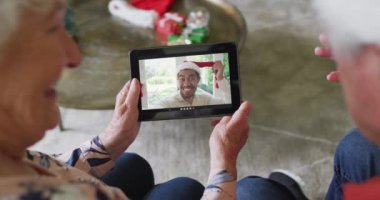 Gülümseyen Kafkasyalı son sınıf çifti, ekrandaki adamla noel videosu için tablet kullanıyor. Noel, şenlik ve iletişim teknolojisi.