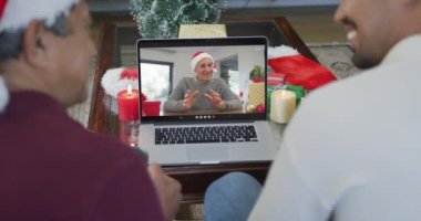 Noel Baba şapkalı iki ırklı baba ve oğul, ekrandaki adamla yılbaşı görüntülerinde dizüstü bilgisayar kullanıyorlar. Noel, şenlik ve iletişim teknolojisi.