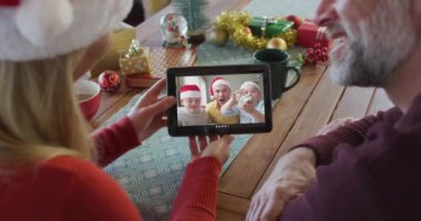 Noel Baba şapkalı, gülümseyen beyaz bir çift ekranda aileleriyle birlikte noel videosu için tablet kullanıyorlar. Noel, şenlik ve iletişim teknolojisi.