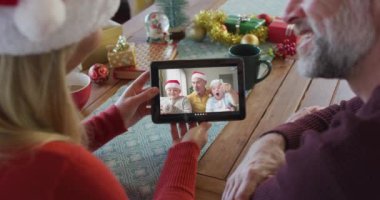 Noel Baba şapkalı, gülümseyen beyaz bir çift ekranda aileleriyle birlikte noel videosu için tablet kullanıyorlar. Noel, şenlik ve iletişim teknolojisi.
