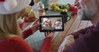 Noel Baba şapkalı, gülümseyen beyaz bir çift ekranda aileleriyle birlikte noel videosu için tablet kullanıyorlar. Noel, şenlik ve iletişim teknolojisi.