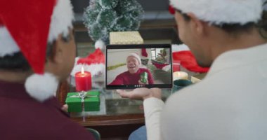 Noel Baba şapkalı iki ırklı baba ve oğul. Ekrandaki adamla noel videosu için tablet kullanıyorlar. Noel, şenlik ve iletişim teknolojisi.