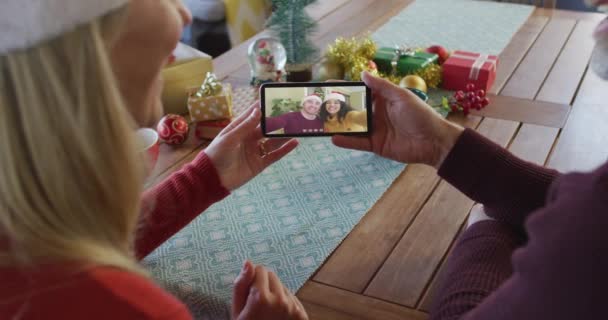 白种人夫妇与桑塔帽使用智能手机与夫妇在屏幕上的圣诞视频通话 圣诞节 节日和通信技术 — 图库视频影像