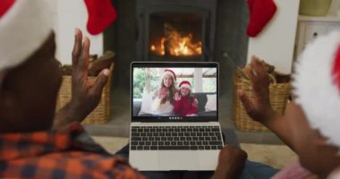 Noel Baba şapkalı Afro-Amerikalı çift, ekranda aileleriyle yılbaşı görüntüsü için dizüstü bilgisayar kullanıyor. Noel, şenlik ve iletişim teknolojisi.