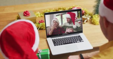 Noel Baba şapkalı beyaz bir baba ve oğul, ekranda aileleriyle yılbaşı görüntülerinde dizüstü bilgisayar kullanıyorlar. Noel, şenlik ve iletişim teknolojisi.