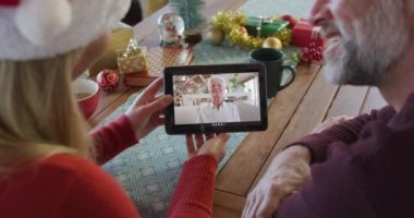 Noel Baba şapkalı beyaz bir çift, ekranda kadınla noel videosu için tablet kullanıyor. Noel, şenlik ve iletişim teknolojisi.