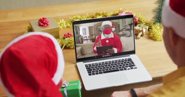 スクリーン上のサンタとクリスマスのビデオ通話のためのラップトップを使用してサンタの帽子を持つ白人の父と息子 19大流行期のクリスマスやお祭りや通信技術は — ストック動画