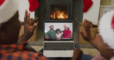 Noel Baba şapkalı Afro-Amerikalı çift, ekranda aileleriyle yılbaşı görüntüsü için dizüstü bilgisayar kullanıyor. Noel, şenlik ve iletişim teknolojisi.