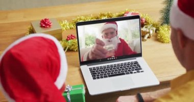 Noel Baba şapkalı beyaz bir baba ve oğul, Noel Baba 'nın ekranda olduğu bir videoda dizüstü bilgisayar kullanıyor. Noel, şenlik ve iletişim teknolojisi.