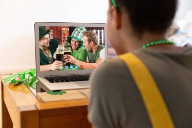 Karışık ırkçı kadın dizüstü bilgisayarında içki içen gülen arkadaşlarına Aziz Patrick Günü videosu çekiyor. Karantina süresince evde tecrit edilmiş İrlandalı koruyucu azizlerin gününü kutluyoruz..