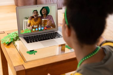 Karışık ırkçı kadın evde dizüstü bilgisayarında bira tutan erkek arkadaşlarıyla Aziz Patrick Günü videosu çekiyor. Karantina süresince evde tecrit edilmiş İrlandalı koruyucu azizlerin gününü kutluyoruz..